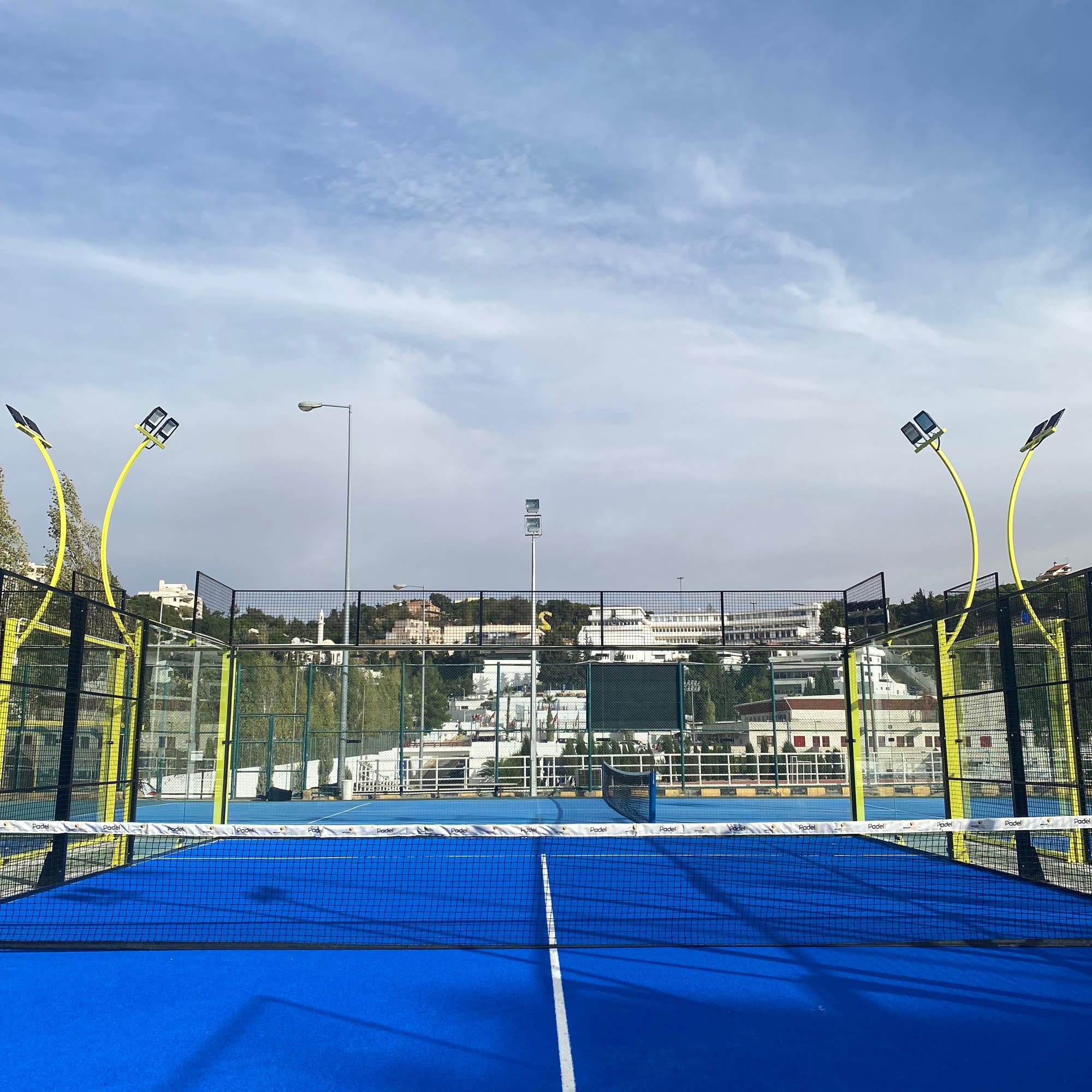 Padel Pro's courts with yellow floodlight posts, seen across the net on a clear day